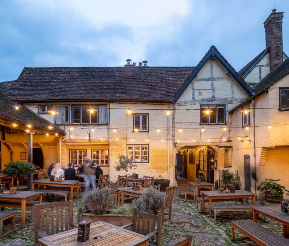 The courtyard of a public house at dusk, with tables and benches, lit by strings of lanterns.