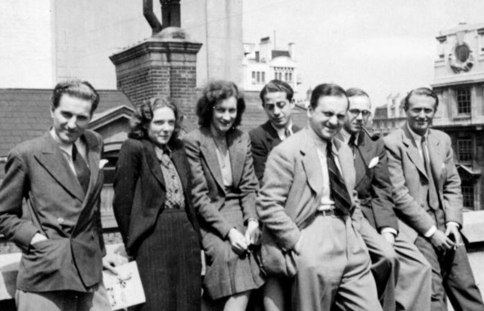 A group of men and women in mid-20th century clothing posing for the camera on a roof space with a cityscape in the background.