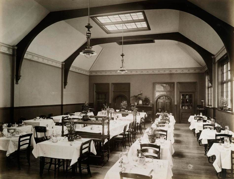 A large Victorian era communal dining room with over twenty tables decorated with tablecloths.
