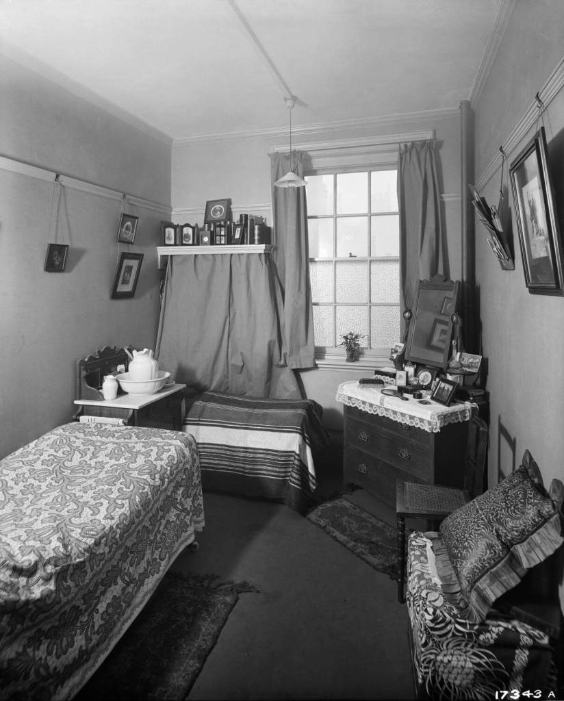 Black and white archive image of the interior of a small room  with a narrow bed, chest of drawers and wash stand.