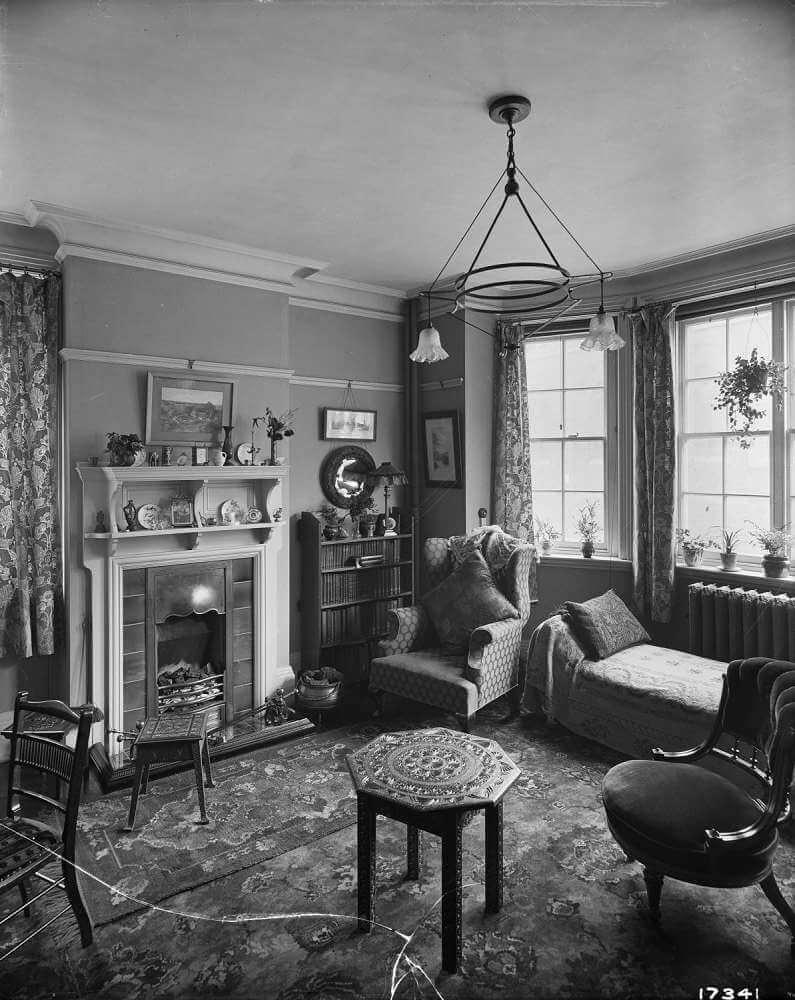 Black and white archive image of an Edwardian room with fireplace, bookcase and a day bed or couch.