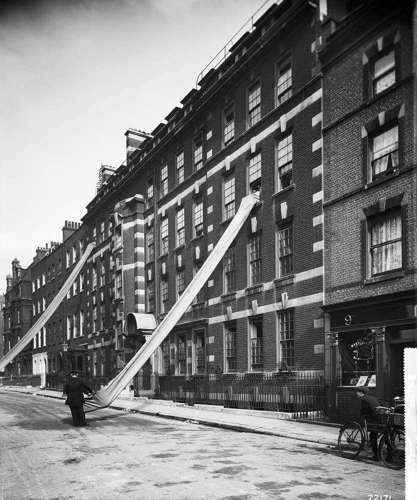 2 long canvas shutes extended from the third floor of a five-storey Victorian building. A uniformed man is holding the end of the shute on the street while a delivery boy looks on.