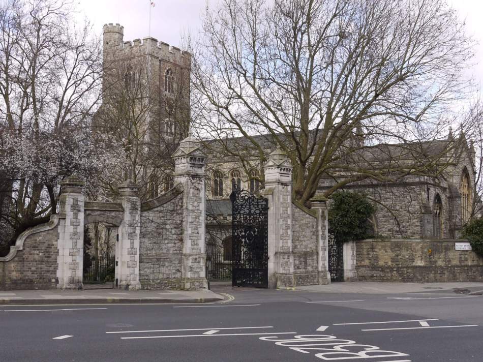 A colour photograph of a church built of stone with surrounding wall and ornate wrought-iron gate.