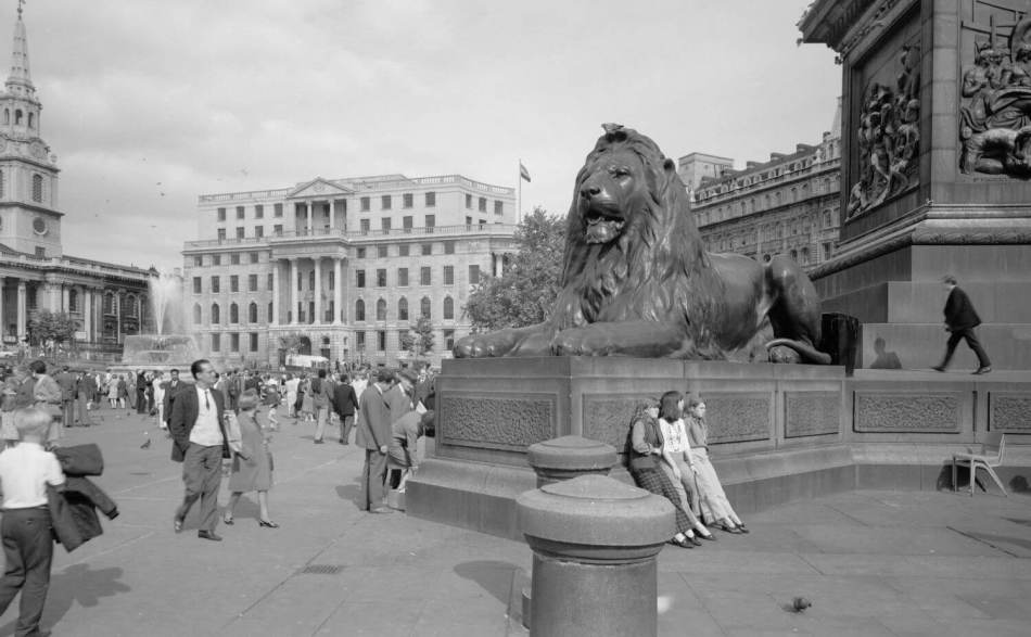 A black and white archive image of the base of a monumental column featuring the sculpture of a lion, with visitors and passers-by.