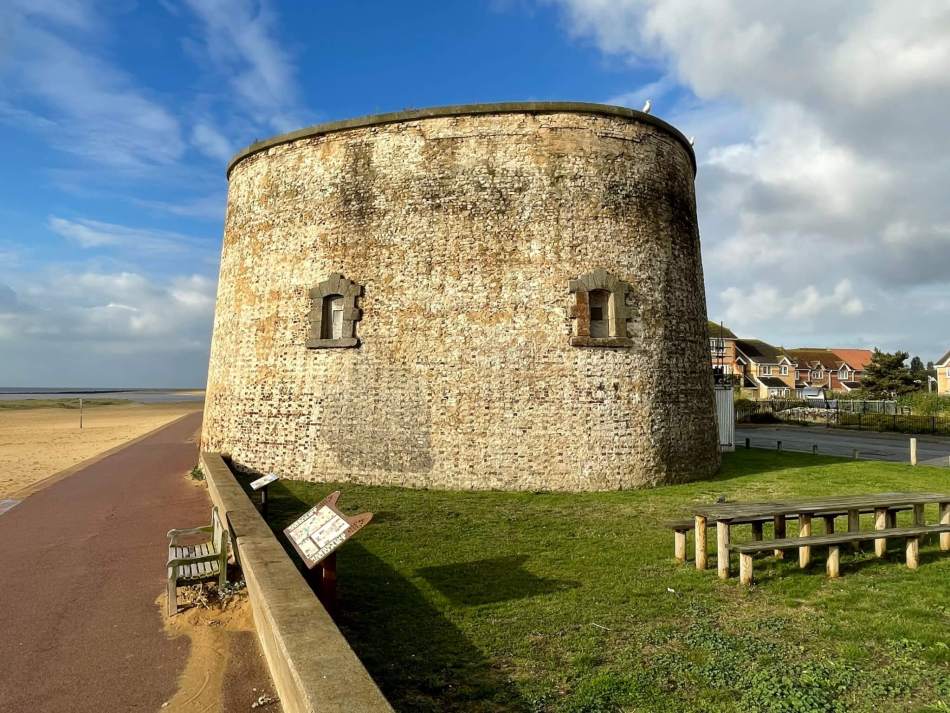 A photograph of a Martello Tower with two windows. Next to the tower is picnic benches on grass. 
