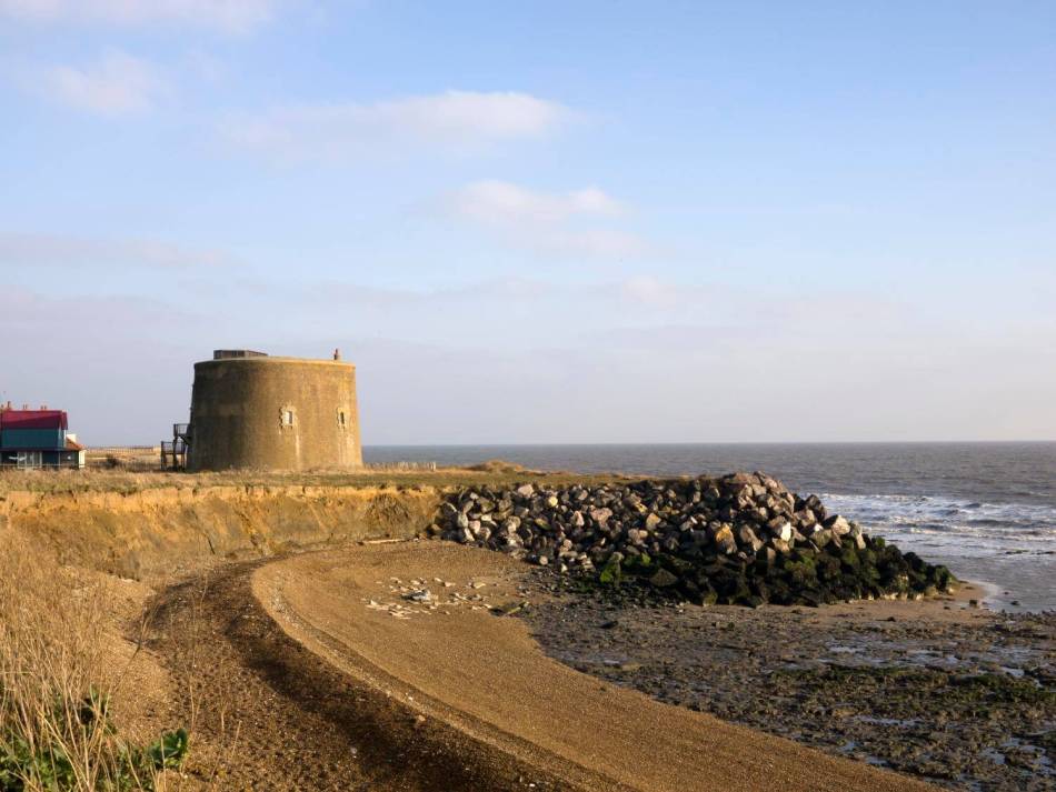 A photograph of a round Martello tower made of stone near the edge of a coastal cliff, overlooking a rocky beach and the sea. 