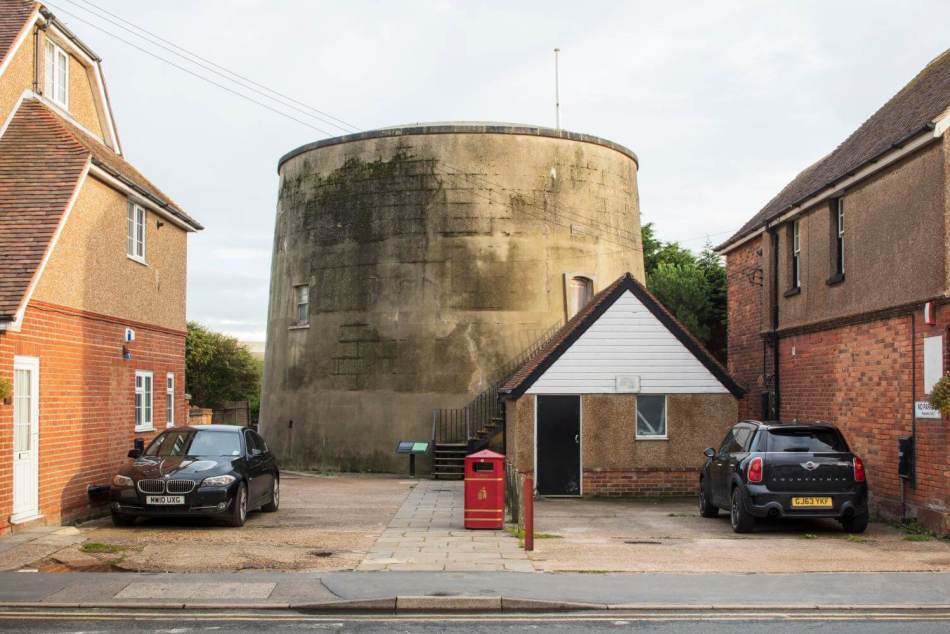A photograph of a large, round, stone structure stands between 2 brick buildings. In the foreground, 2 black cars are parked on either side. 