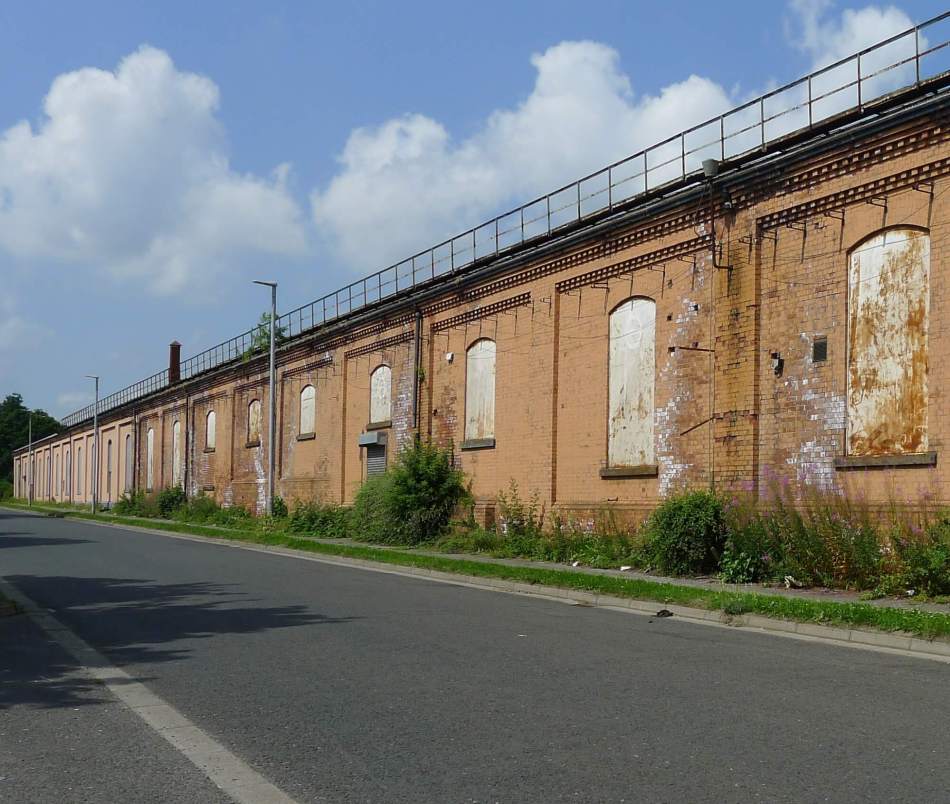 A photograph of the long side of a dilapidated brick wagon works building with blocked up windows with a road running along beside it.