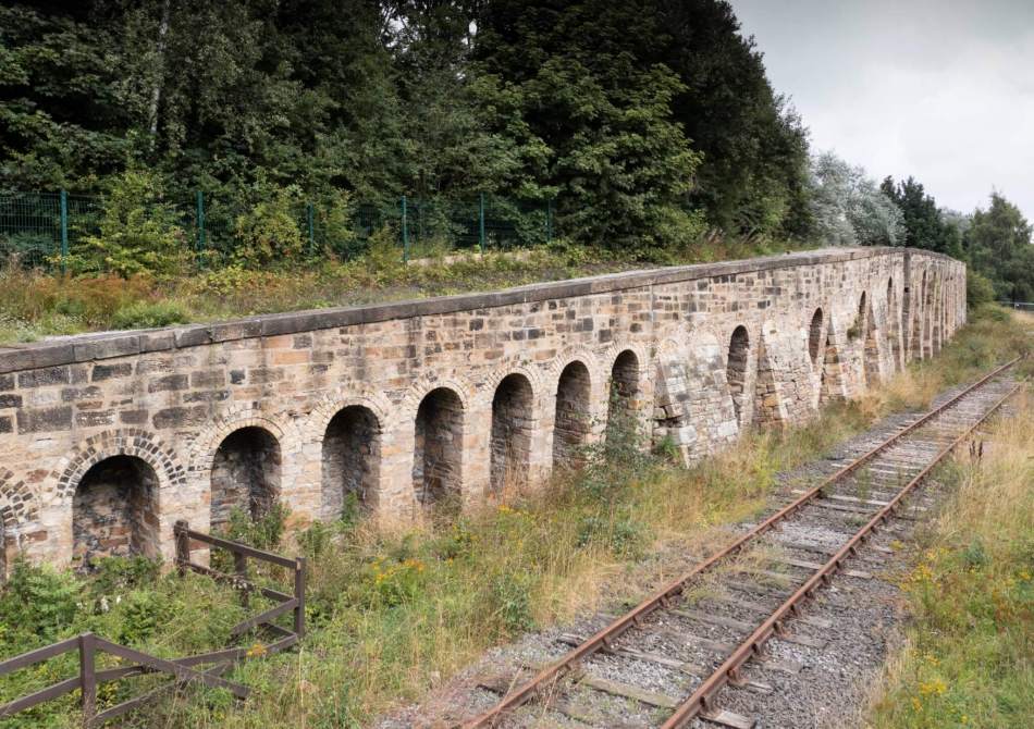 A photograph of a long row of a stone railway coal drops beside an empty railway track. It is surrounded by trees and long grass.