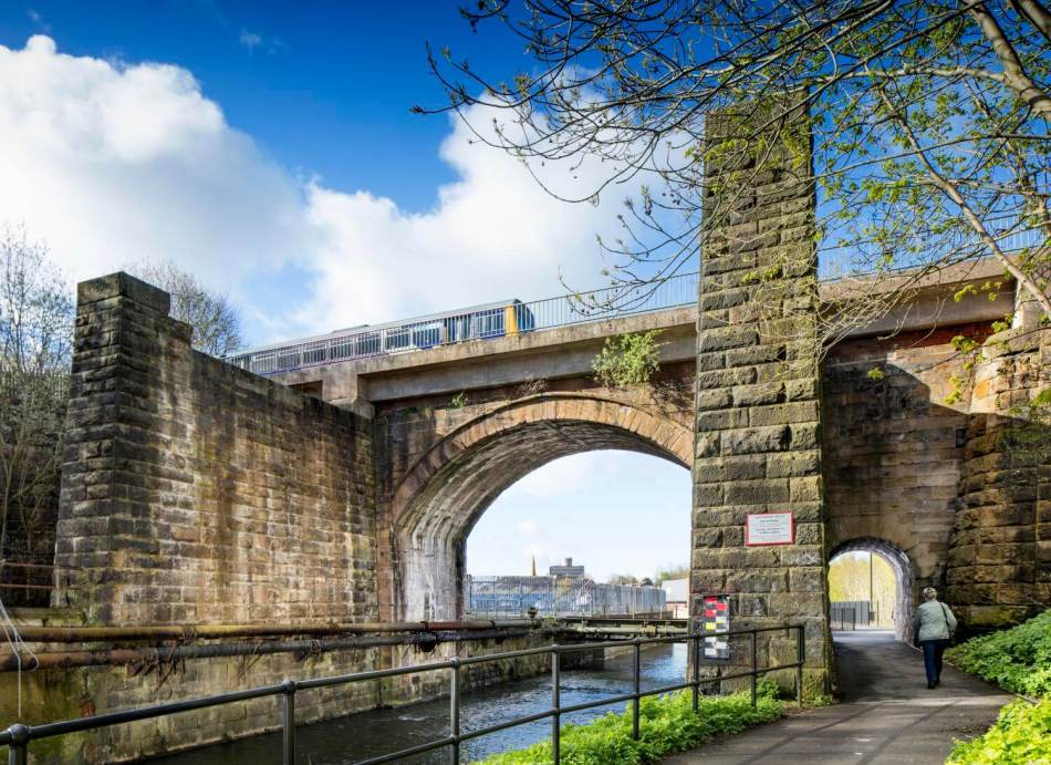A photograph of a stone railway bridge over a small canal. A train travels across the bridge.