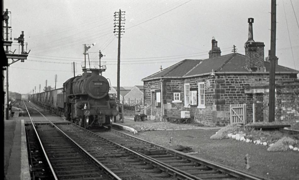 A black and white photograph of a small, single-storey stone railway station beside a railway track. A train travels across the track.