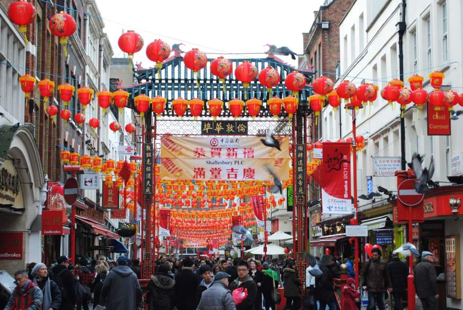 A busy Chinatown street decorated with numerous red lanterns hanging overhead. A large archway adorned with Chinese characters spans the street. Crowds of people walk below, surrounded by red banners and signs.