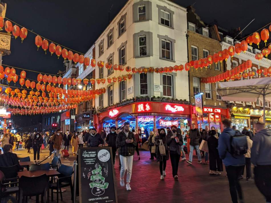 A bustling city street at night is adorned with strings of red lanterns. A crowd of people walk beside illuminated shops and restaurants.
