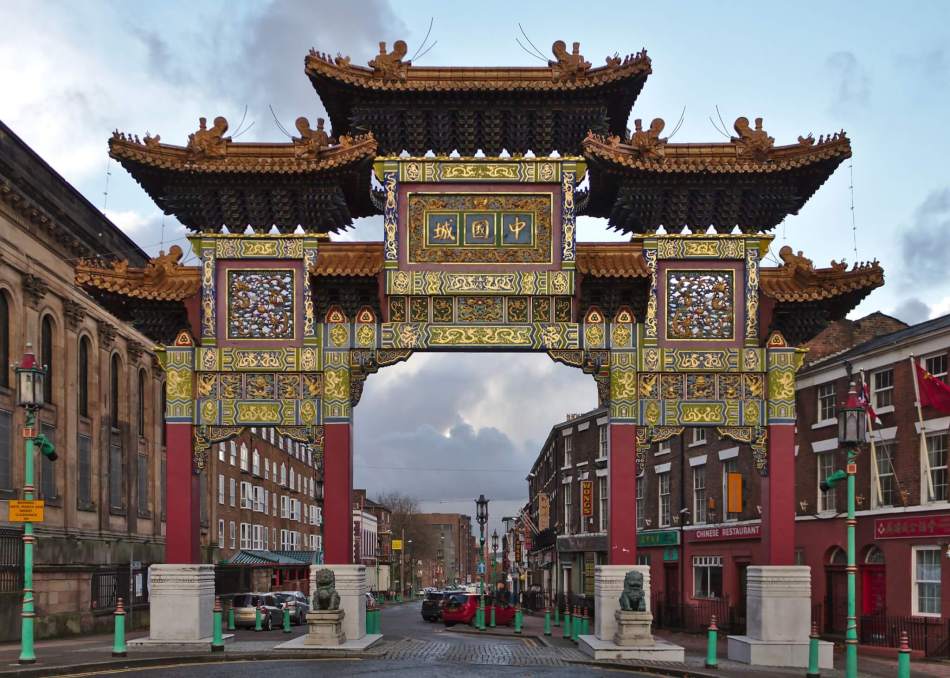 An intricately decorated Chinese archway with traditional green, red, and gold colours, standing at the entrance to a street lined with brick buildings and Chinese businesses. Stone lion statues guard the arch.