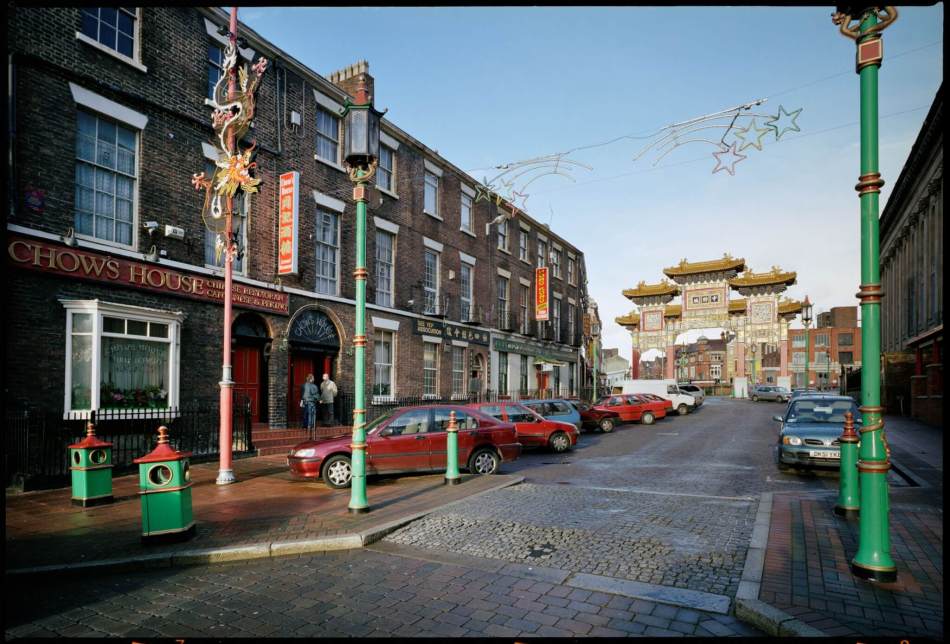 A street scene in a Chinatown area, featuring a prominent Chinese archway adorned with intricate patterns in the background. The street is lined with brick buildings housing Chinese restaurants and shops. Several bright green and red lampposts and decorative elements hang overhead.
