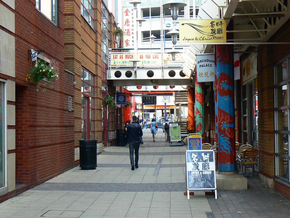 A narrow urban alley lined with brick buildings and decorated pillars painted with vibrant patterns. The alley features hanging flower baskets and various restaurant signs.
