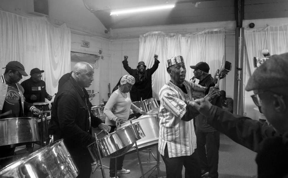 A black and white photograph of a lively group of musicians plays steel drums in a rehearsal space,