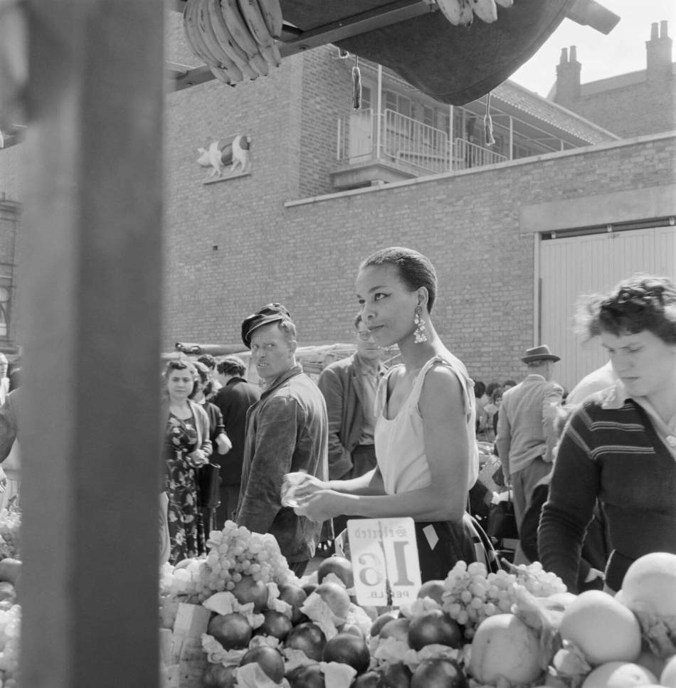 A black and white photograph of a market with a woman examining produce, surrounded by various fruits and vegetables. Nearby, a man looks on, while others engage in shopping and conversation.
