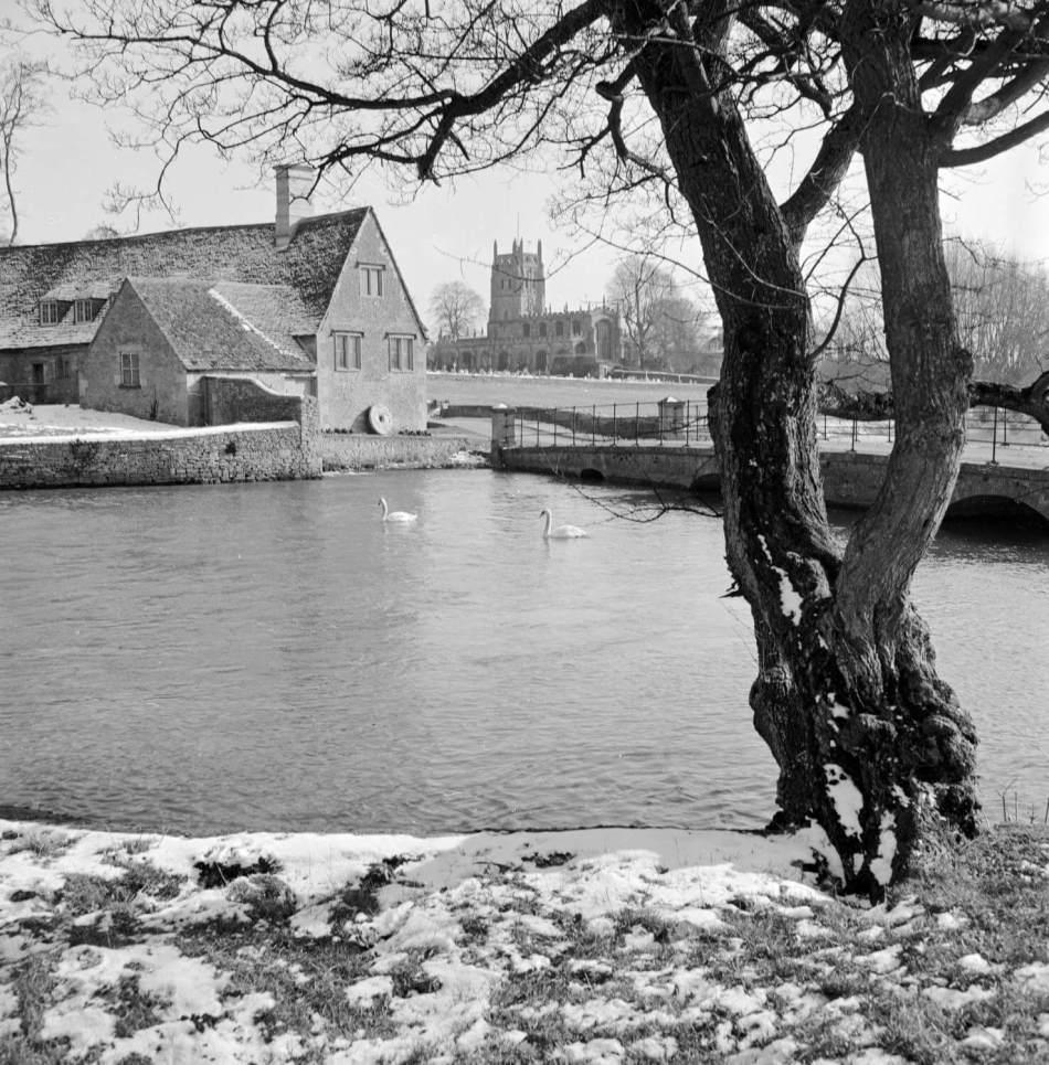 A black and white photograph of a lake with swans swimming. In the background is a mill and a church. 