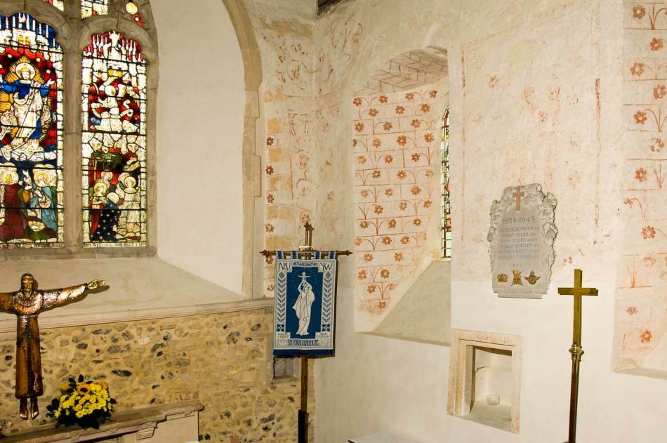 A photograph of the interior of a church chancel showing floral wall paintings around the stained glass windows.