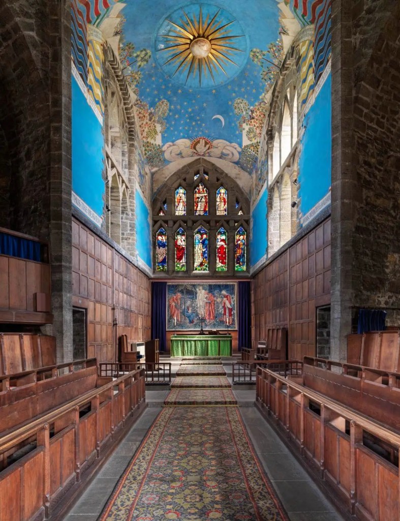 A photograph of the interior of the choir of a church featuring wooden panelled seating. The ceiling is covered in colourful, detailed wall paintings, including a large sun and stars.