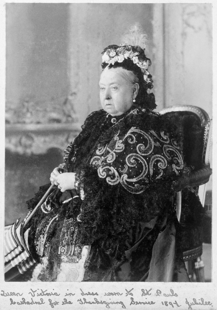 A black and white photograph of Queen Victoria seated, adorned in elaborate attire with a floral headdress.