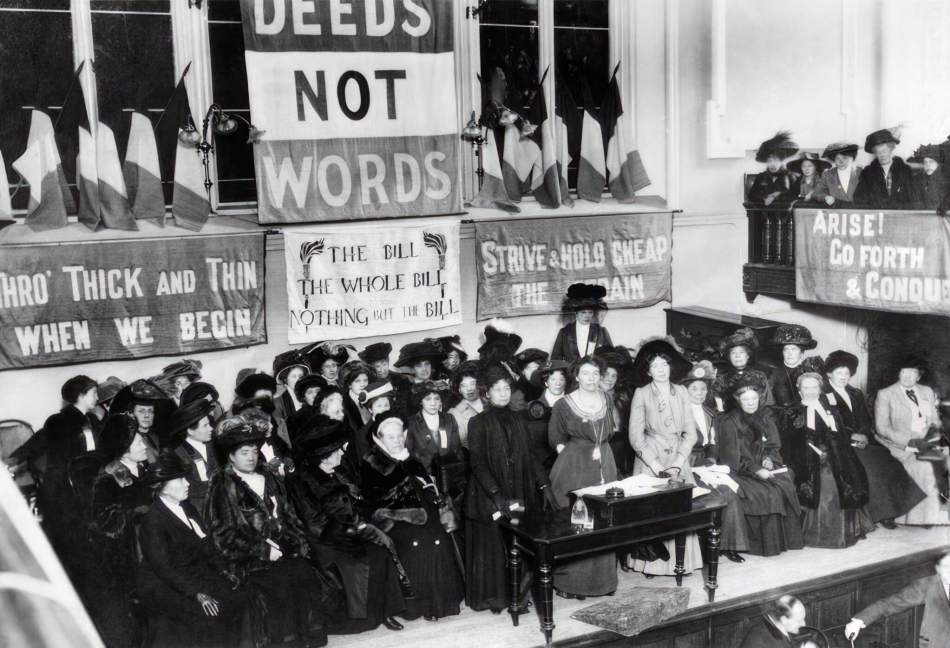 A black and white photograph of a group of women in Edwardian dress standing on a stage surrounded by women's rights campaign banners featuring wording such as 'Deeds Not Words' and 'Arise! Go forth and conquer.'