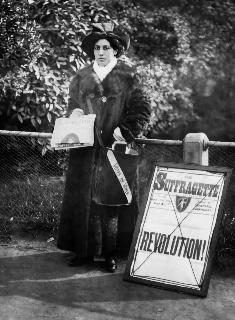 A black and white photograph of Sophia Duleep Singh standing in a park and holding copies of a newspaper. The bag she carries has the words 'votes for women' on it.