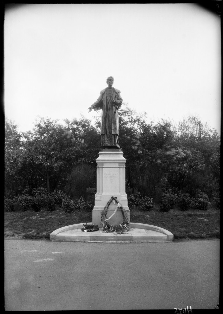 A black and white photograph of a statue of Emmeline Pankhurst on a stone plinth in a park.