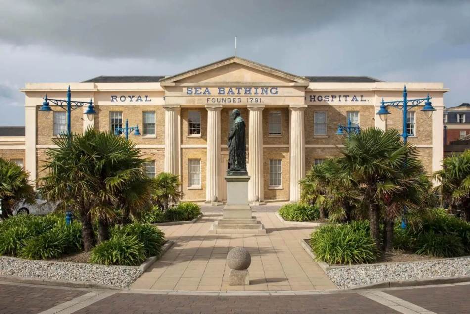 A photograph of the exterior of a large, Greek Revival-style hospital building with tall columns around the centre of the building. Gardens filled with palm trees, a statue and pathways feature in front of the building.