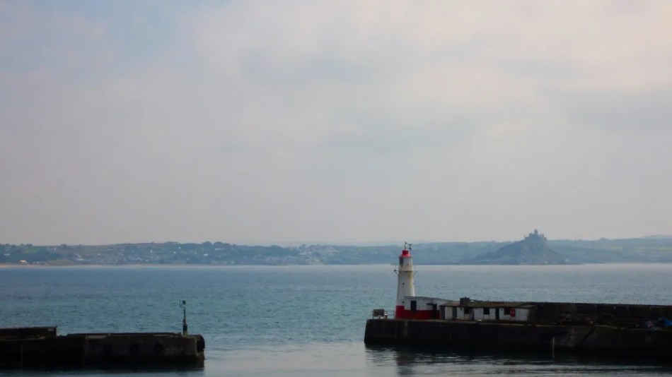 A photograph of a small lighthouse beside a small, single-storey, two-tone building on the end of a pier jutting out into the sea.