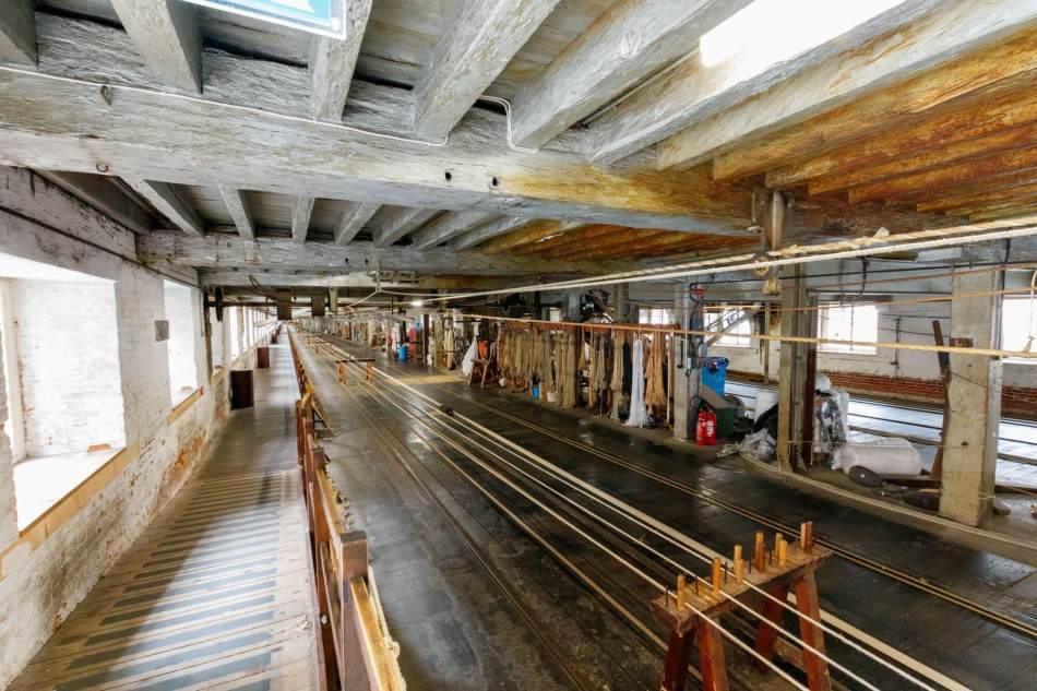 A photograph of the interior of a large, long manufacturing building with low beams on the ceiling. A wooden spinning machine is at the centre, with long sections of rope strung out tightly between each machine.