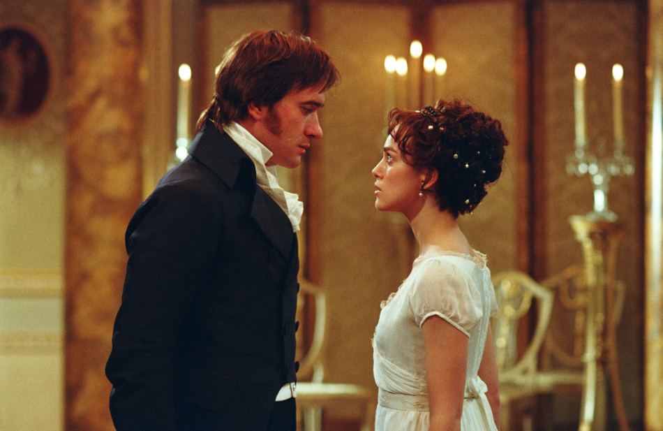 A photograph of Matthew Macfadyen and Keira Knightley dressed in early 19th-century costumes, standing in grand room, staring at one another.