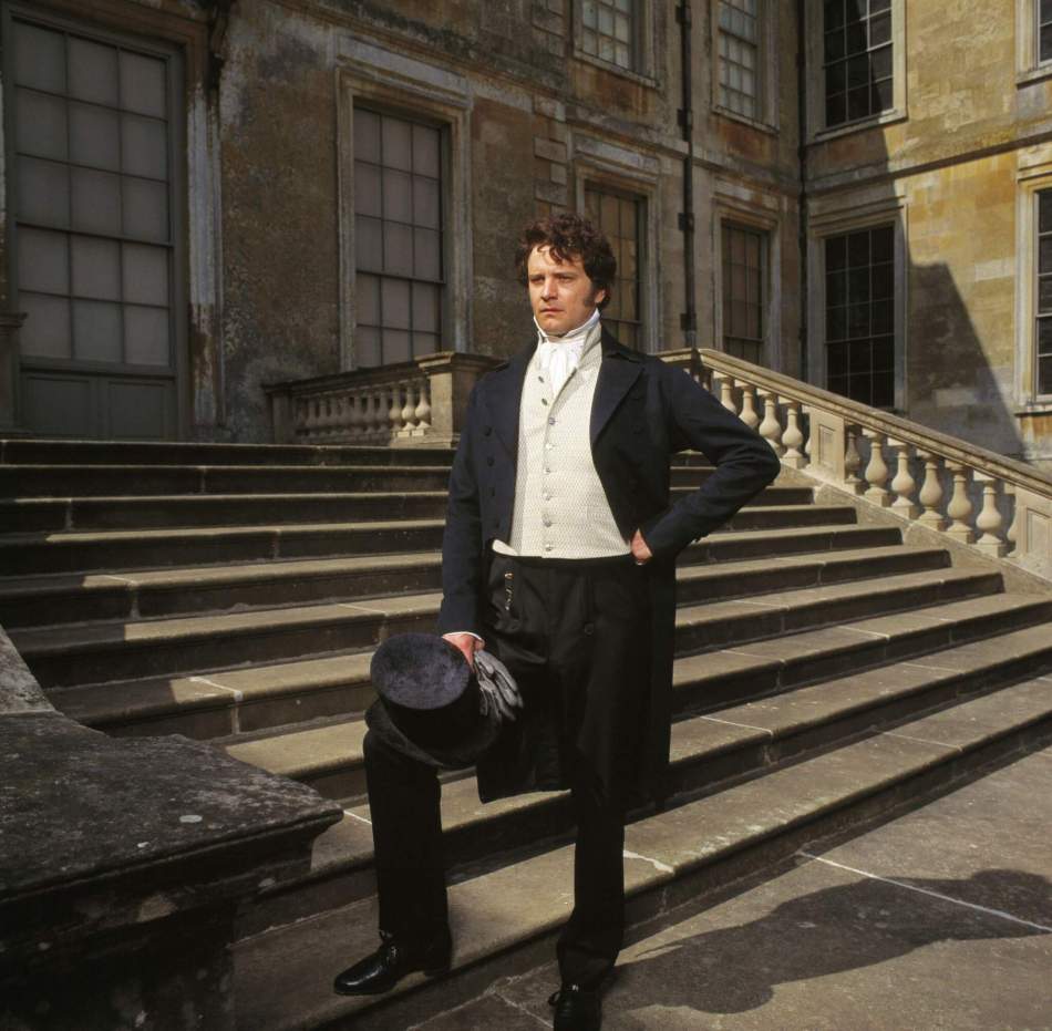 A photograph of Colin Firth dressed in an early 19th-century suit, standing on the steps of a grand house.