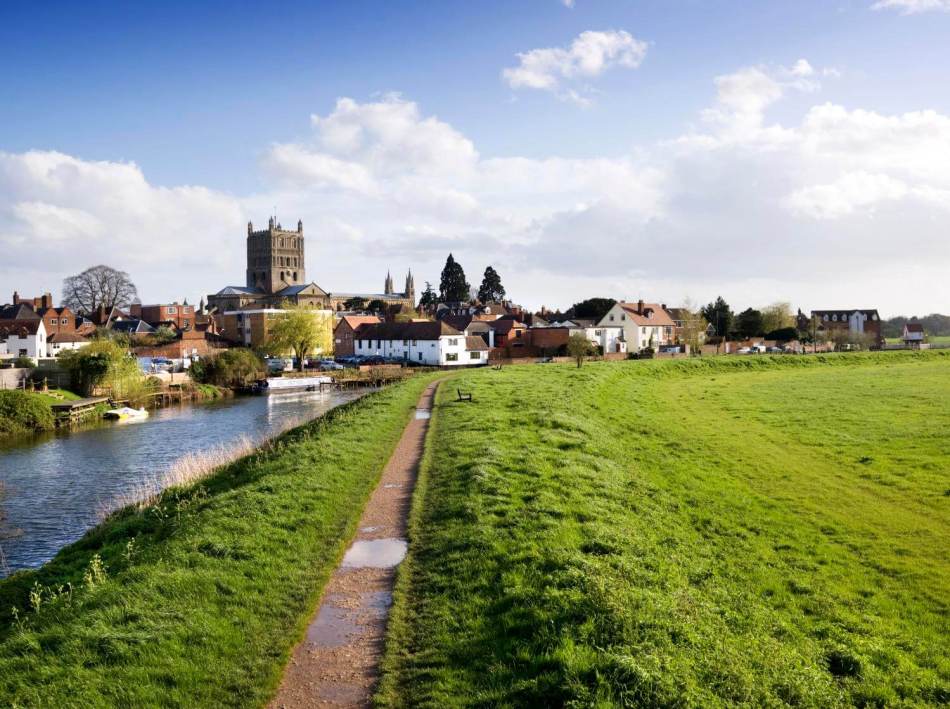 A photograph of a common green with a river to the left, with an abbey tower and multiple historic buildings in the background.