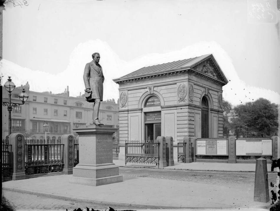 A black and white photograph of a statue of Robert Stephenson on a plinth beside a small classical-style building.