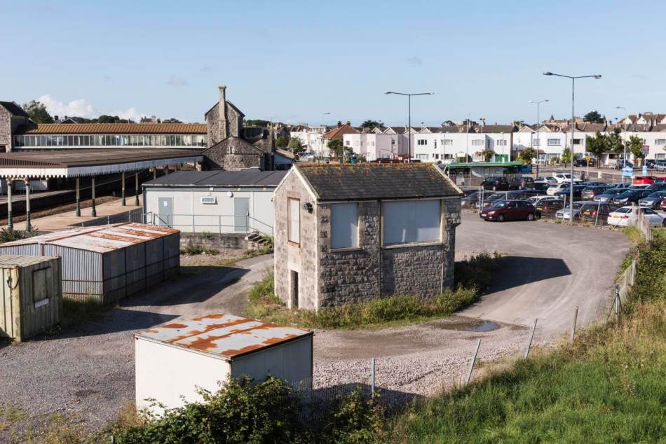 A photograph of a small signal box building with boarded-up windows. A railway platform is in the background.