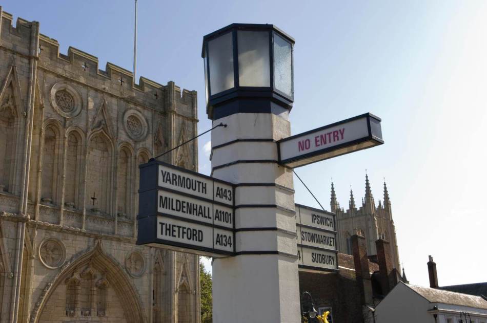 A photograph of a road sign featuring 6 different locations with a small lamp on the top of it. A cathedral can be seen in the background.