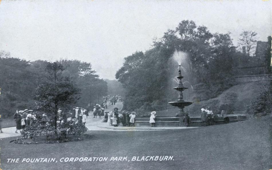 A black and white photograph on a postcard showing children leaning over the edge of a large stone water fountain in a park.