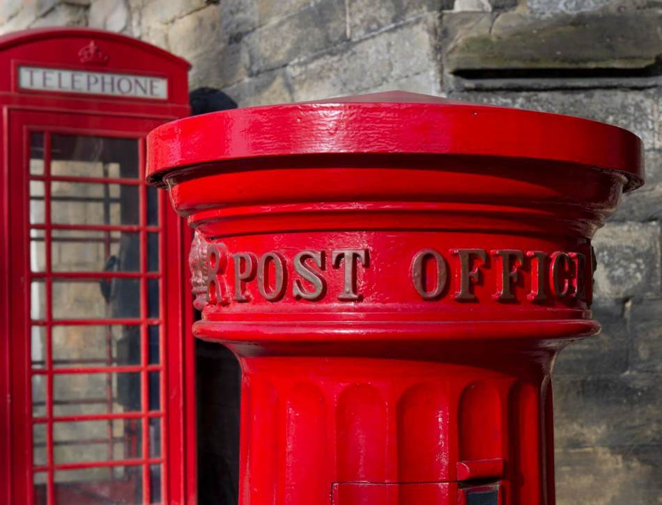 A photograph of a close up of the top of a post box featuring the wording 'Post Office'. A telephone box can be seen in the background.