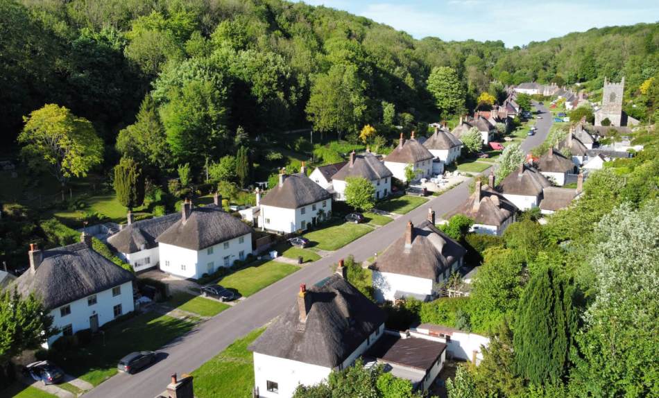 An aerial photograph of rows of semi-detached thatched cottages on a road surrounded by woodland.