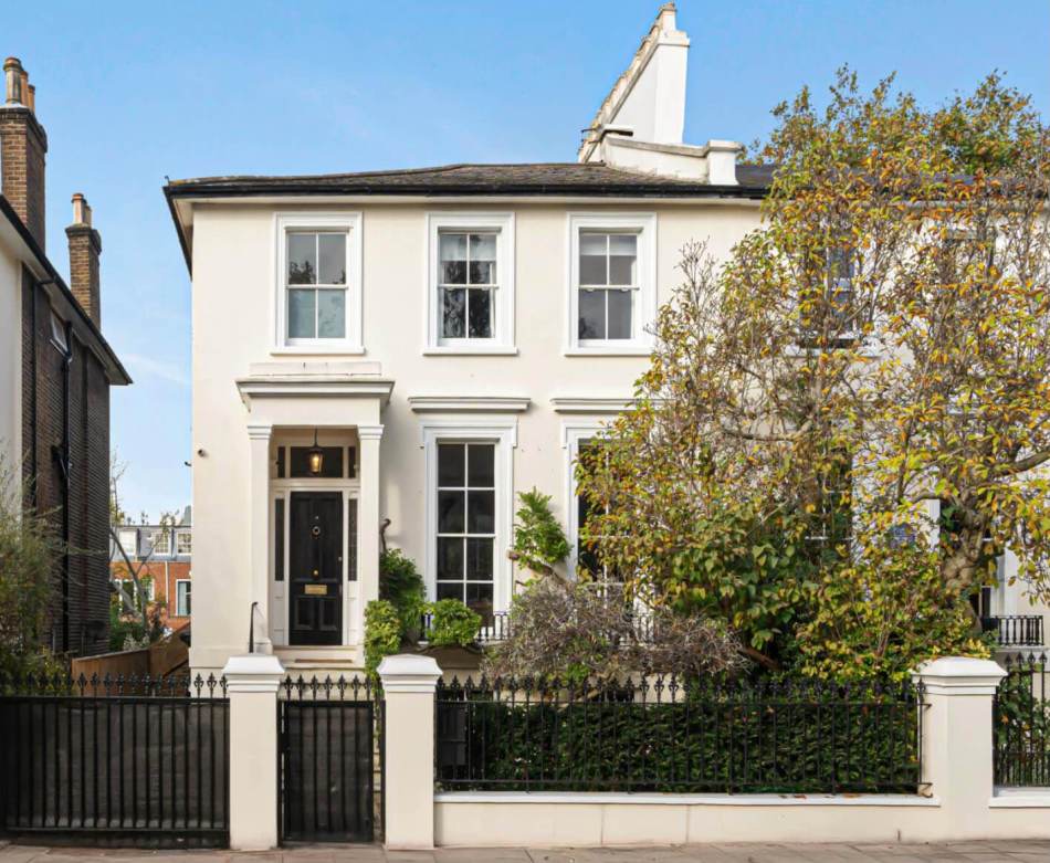 A photograph of the exterior of a 2-storey semi-detached stuccoed Italianate house surrounded by a metal fence and bushes.