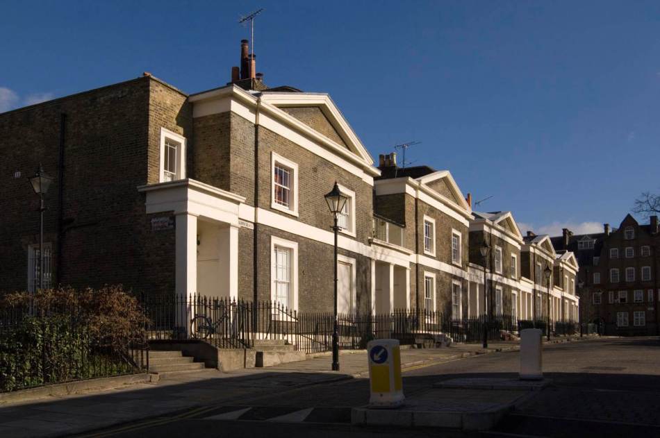 A photograph of the exterior of a row of grand semi-detached linked houses with sash windows beside a road.