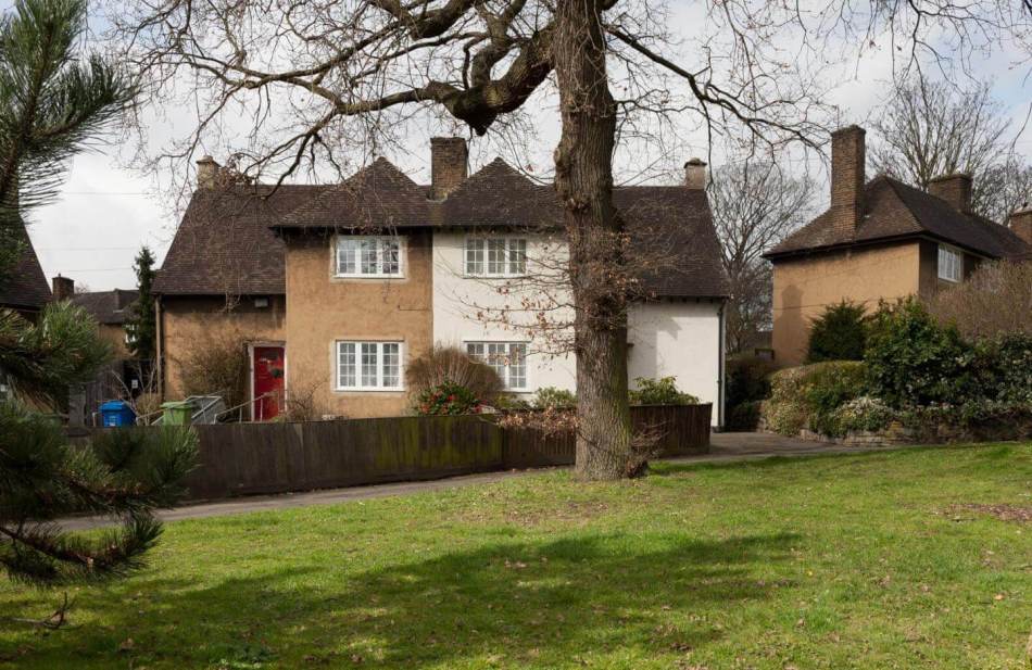A photograph of the exterior of 2 semi-detached houses with a large green space and tree in front.
