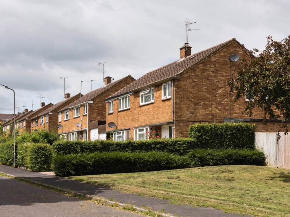 A photograph of a row of post-war semi-detached houses on a road with front gardens and large bushes.