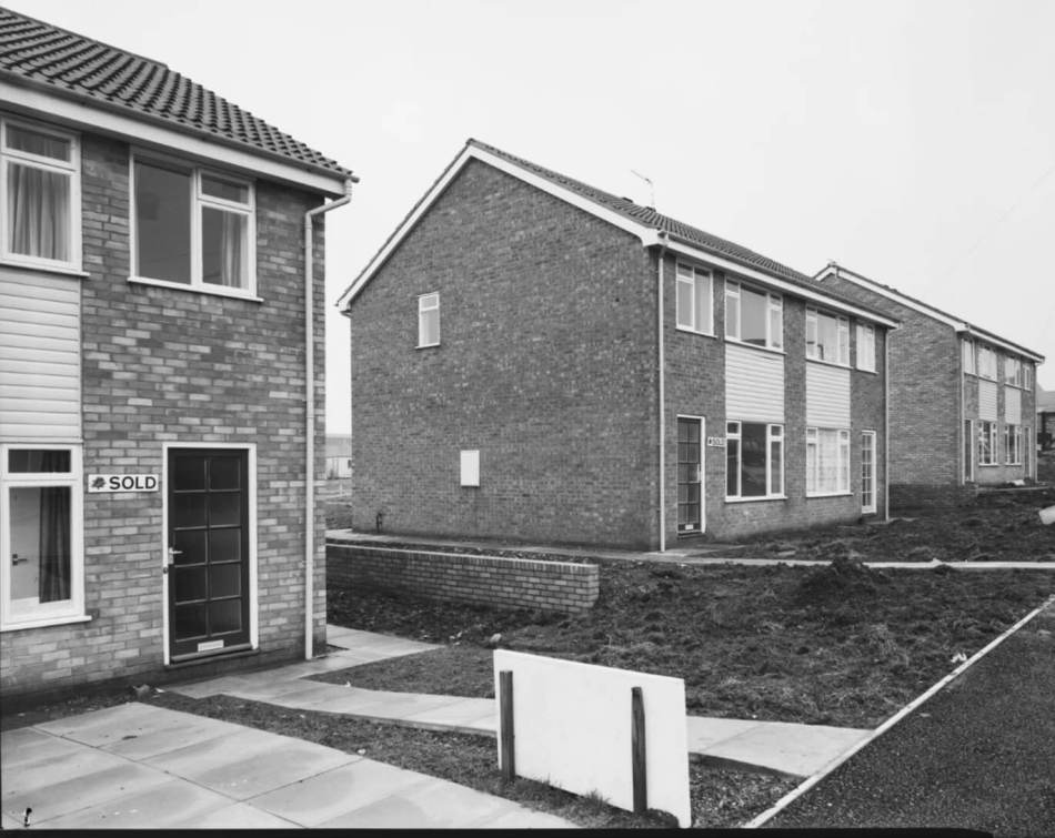 A black and white photograph of newly built semi-detached houses with 'sold' signs by the front doors.