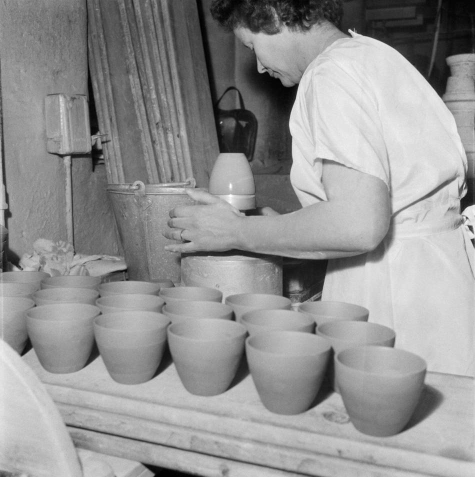 A black and white photograph of a woman smoothing the sides of a small pot on a stand. Rows of small pots are laid out on a bench beside her.