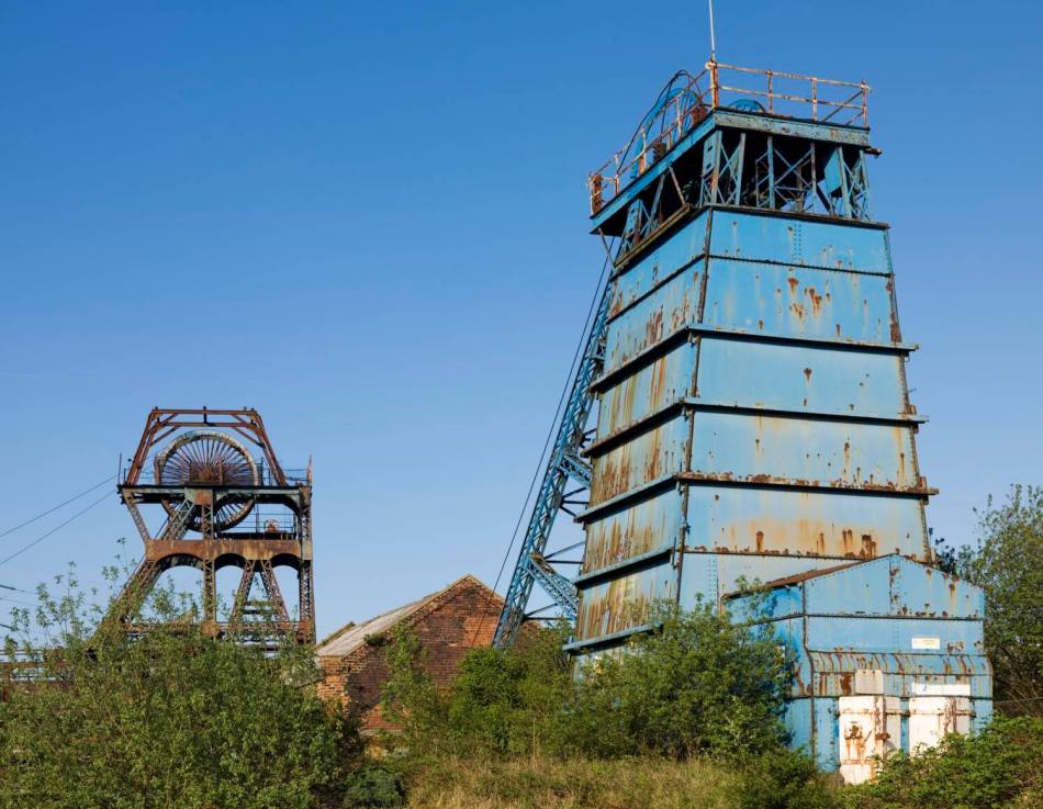 A photograph of 2 sets of headgear on a disused colliery site surrounded by trees and overgrown plants.