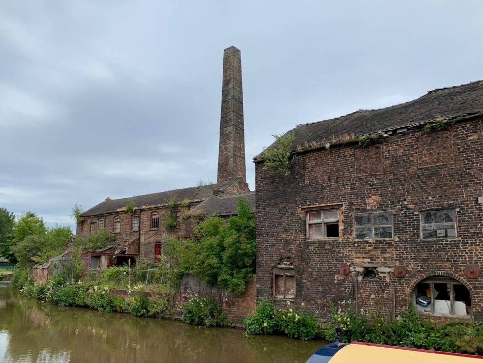 A photograph of the exterior of a derelict pottery building with a tall chimney and warehouse beside a canal.