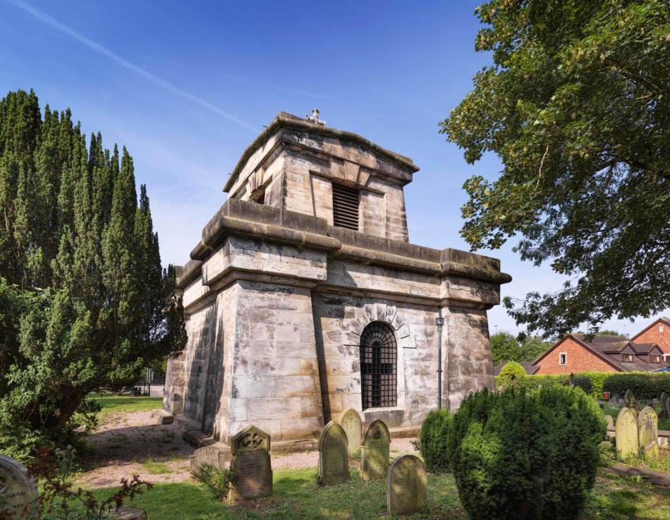 A photograph of the exterior of a stone mausoleum in a cemetery surrounded by gravestones.